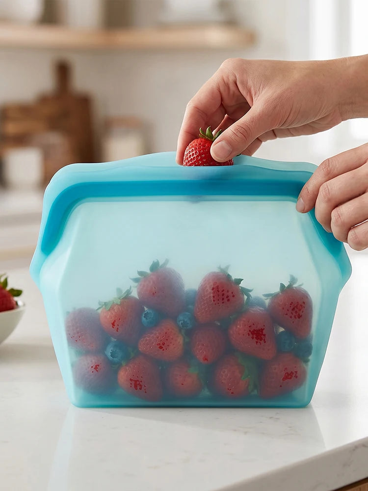 A hand is placing a strawberry into a light blue silicone storage bag filled with fresh strawberries and blueberries.