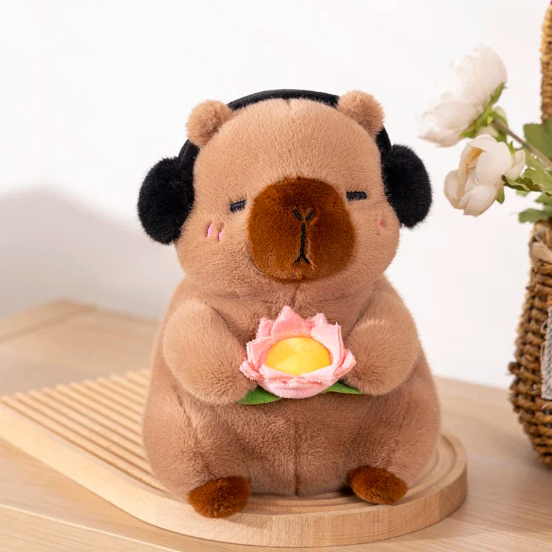 A soft brown capybara plush toy is sitting and holding a pink lotus flower on a wooden surface.