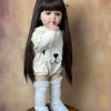 A lifelike 55 cm soft silicone baby doll with long dark hair wearing a white outfit and knee socks.
