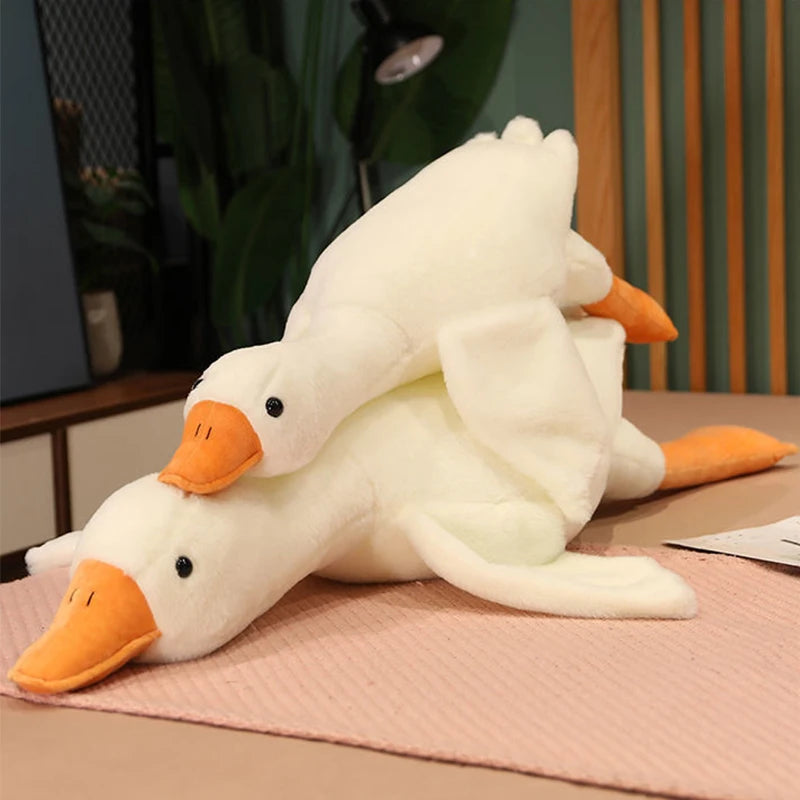 A white plush goose pillow with orange beak and feet lying on a pink surface.