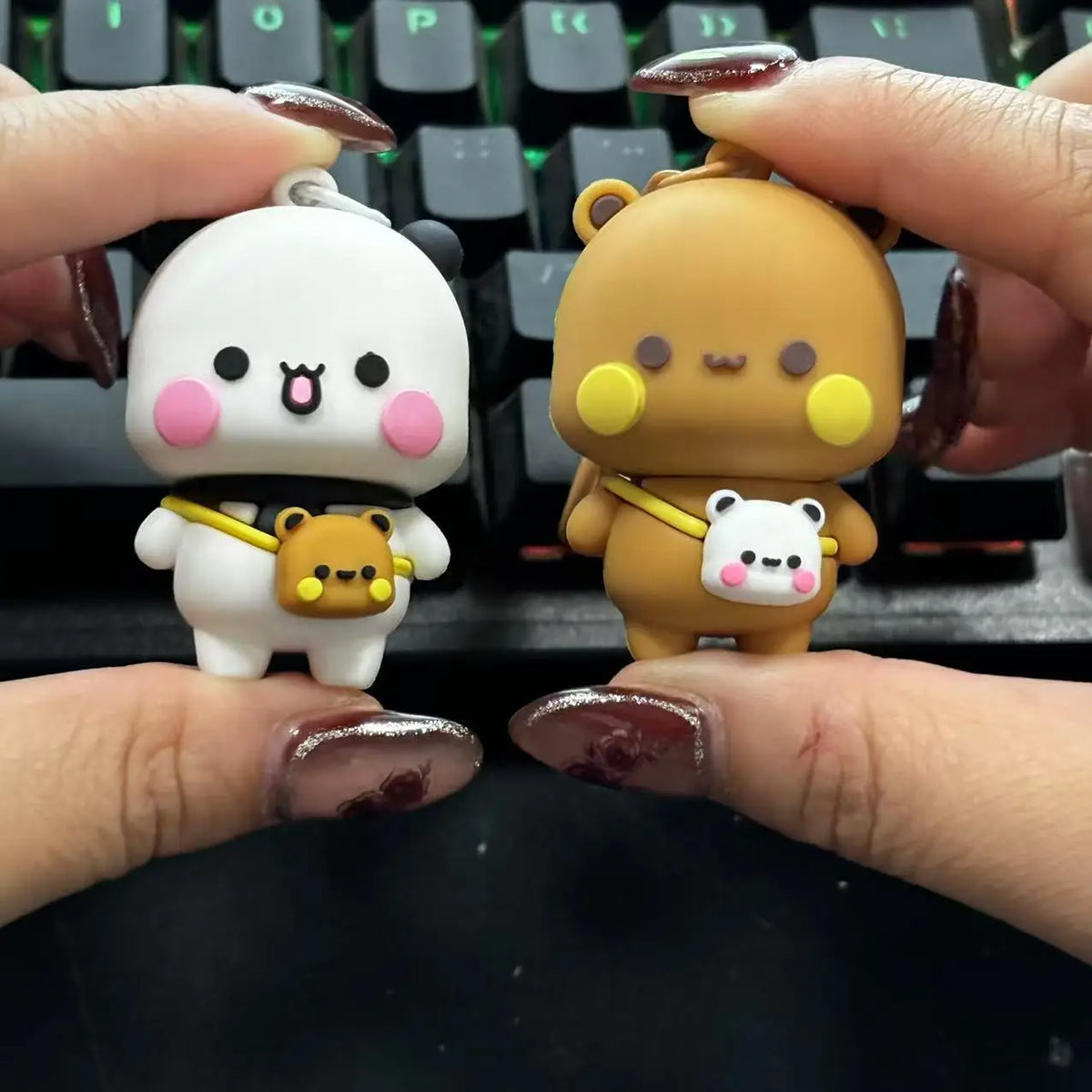 A pair of cute cartoon bear keychains, one white and pink, and one brown, are held between fingers above a keyboard.