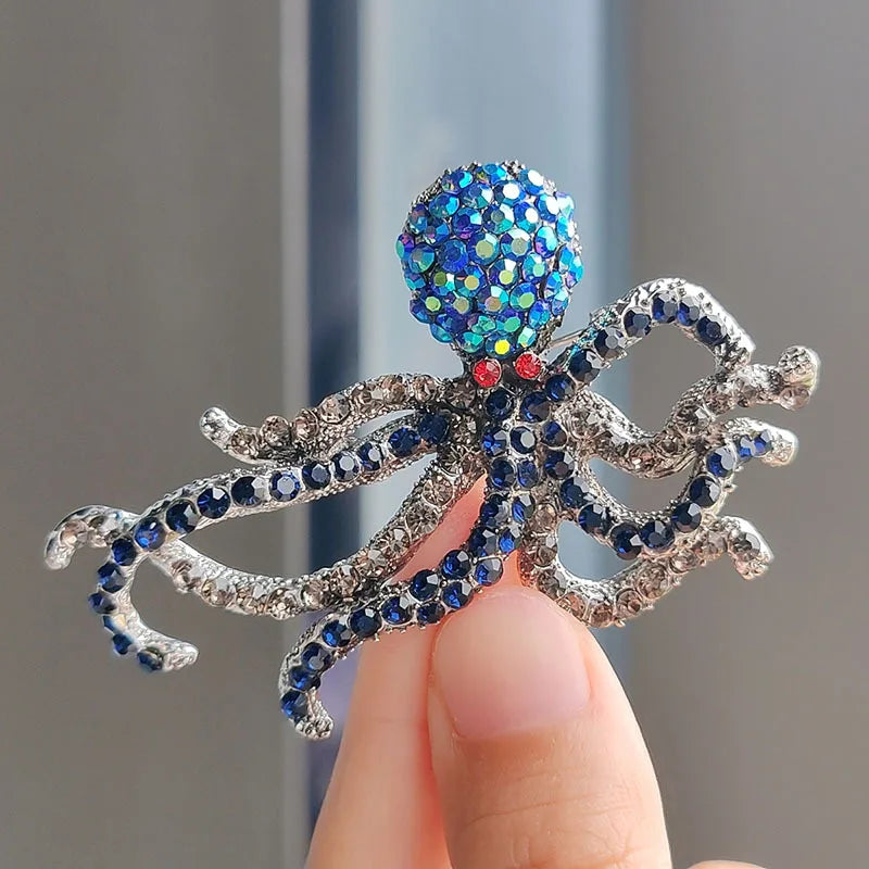 A close-up of a rhinestone turtle brooch pin with iridescent blue and teal stones being held by a hand.