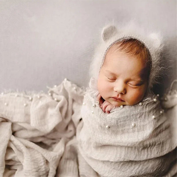A newborn baby sleeping wrapped in a light knit fabric with a pearl headband.