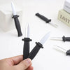 A hand holds a black retractable realistic fake knife prank toy with silver blades on a white surface.