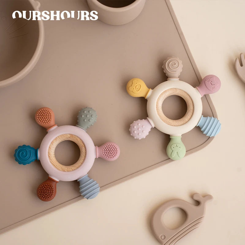 A collection of colorful, rudder-shaped silicone baby teethers with wooden rings arranged on a light surface.