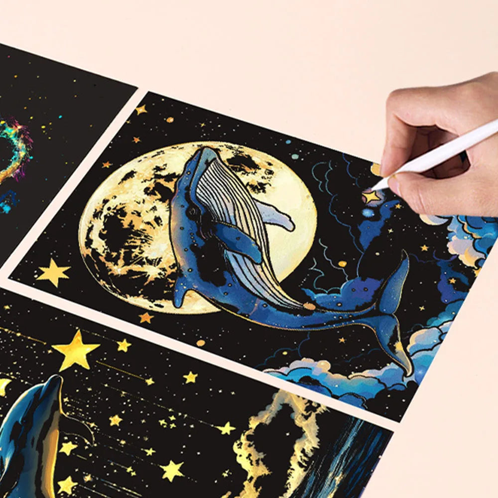 A hand is writing on a piece of glow-in-the-dark scratch art featuring a whale against a moon and starry background.