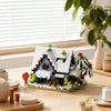 A miniature wooden building set depicting a cabin and windmill scene with greenery on a light wooden surface.