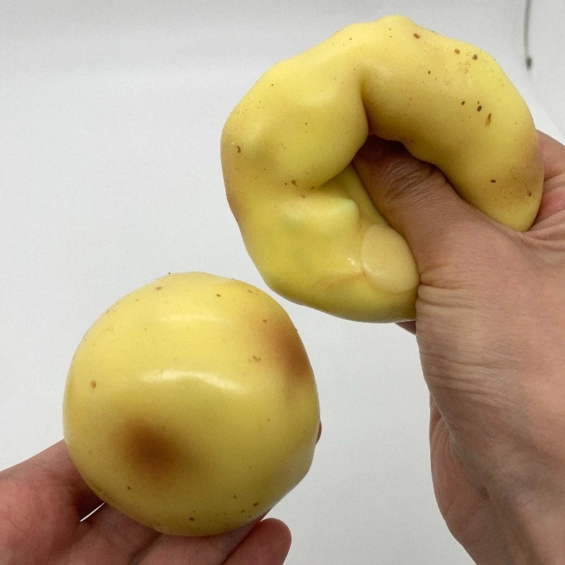 A hand holds a yellow, squishy potato stress ball with brown marks against a white background.