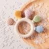 Silikon-Teether-Ring mit mehreren strukturierten Silikonspitzen auf einem Holzbrett, umgeben von kleinen klaren Silikonperlen