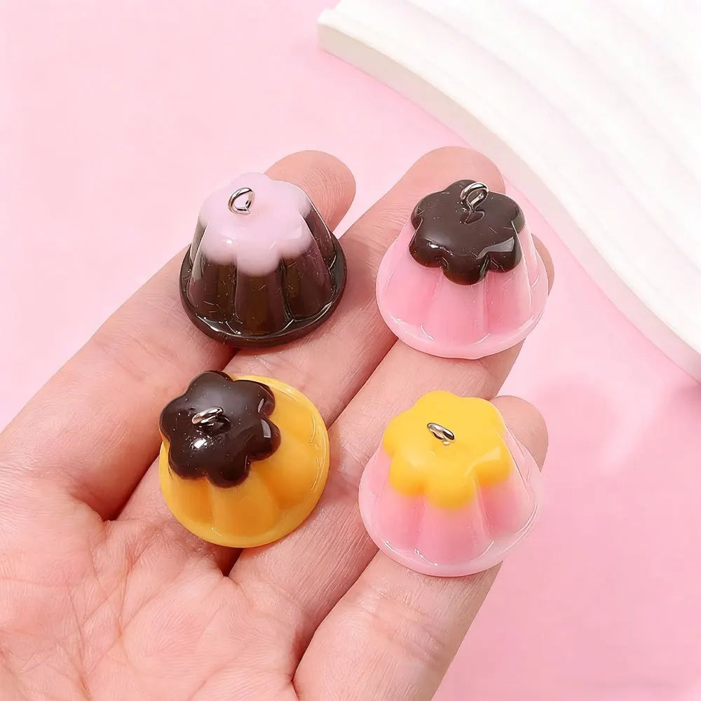 Three pudding-shaped resin pendants in pink, brown, and yellow are displayed on a hand against a light pink background.