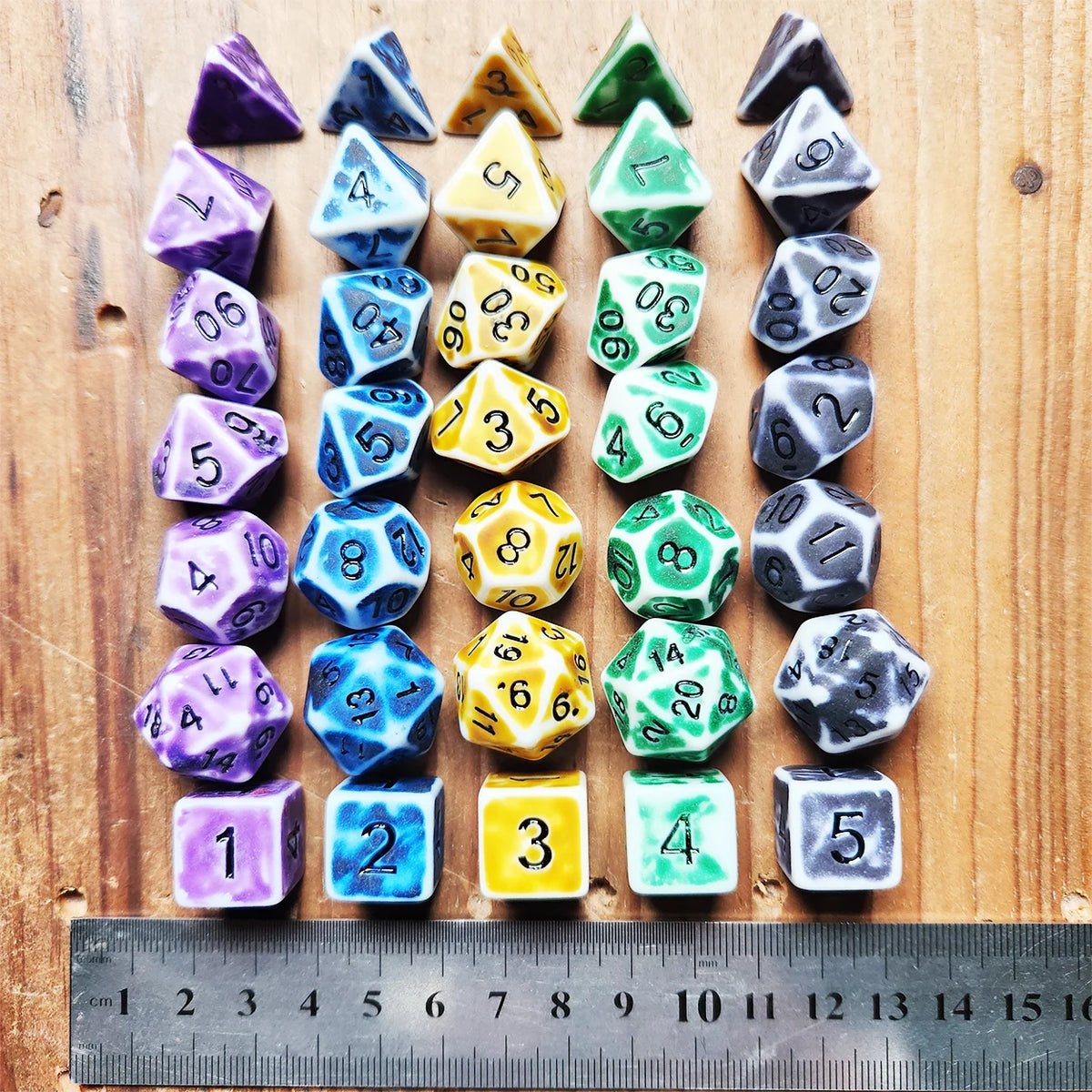 A set of complete polyhedral dice in various colors and shapes laid out on a wooden surface next to a ruler.