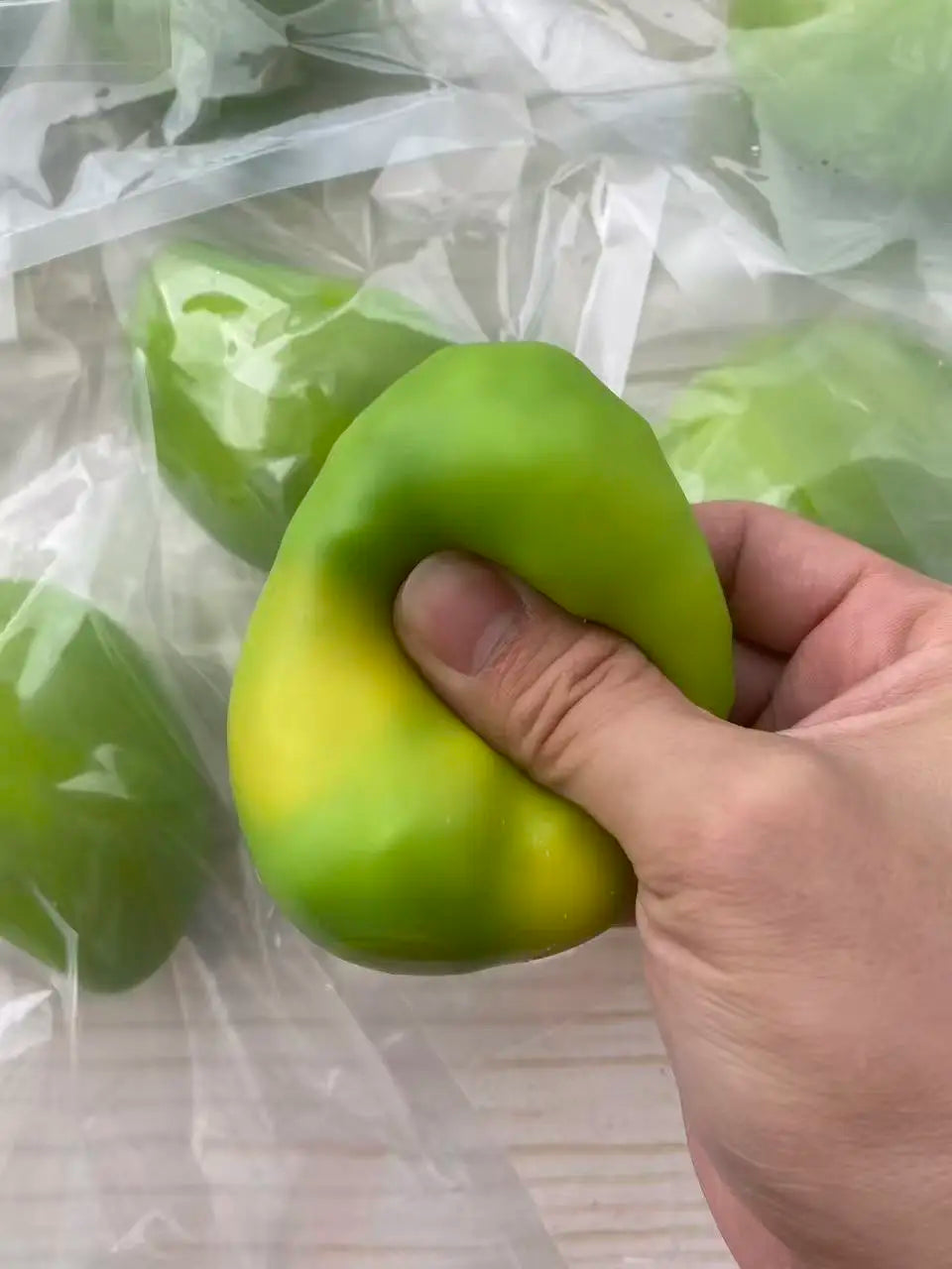 A hand holds a bright green, squishy stress ball with a yellow tint inside clear plastic packaging.