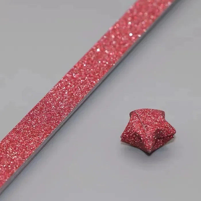 A close-up of a pink glitter paper strip with an origami star shape attached, used for crafting.