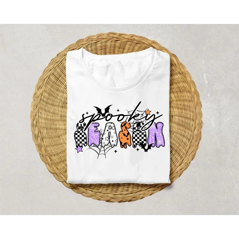 White t-shirt featuring black spooky-themed iron-on patches with purple and orange designs on a woven mat.