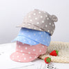 A stack of baby cotton baseball caps in gray, blue, and pink with white polka dots resting on a woven mat.