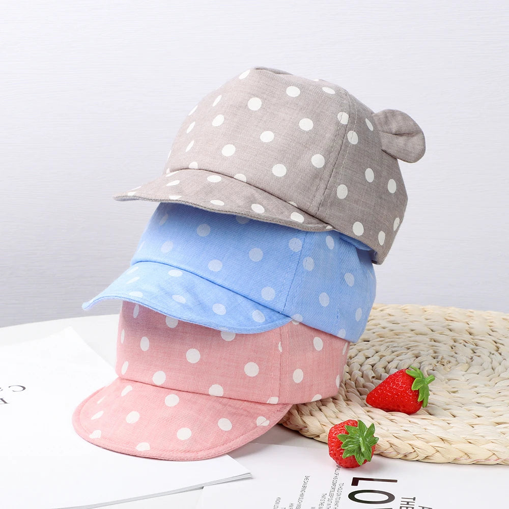 A stack of baby cotton baseball caps in gray, blue, and pink with white polka dots resting on a woven mat.