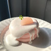 A pink cake with a smooth, white glaze and a small green leaf decoration on a white plate.