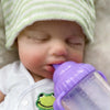 A realistic reborn baby doll wearing a white outfit is shown holding a purple baby bottle.