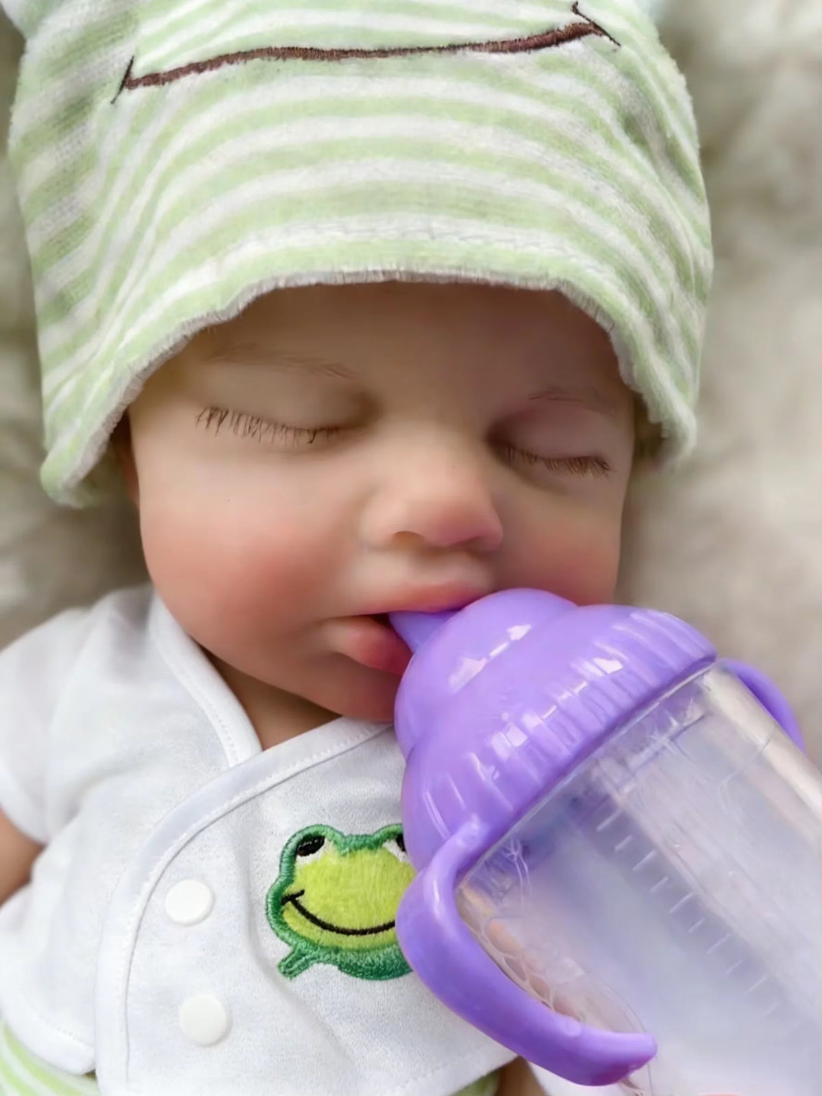 A realistic reborn baby doll wearing a white outfit is shown holding a purple baby bottle.