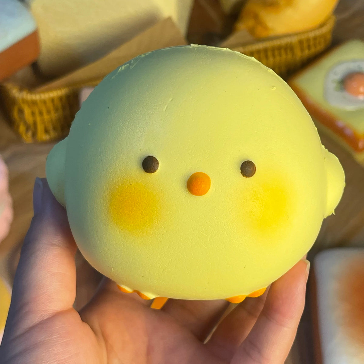 A hand holds a yellow squishy toy shaped like a chick with orange feet and facial features.