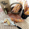A miniature baguette is shown next to a wooden cutting board and a woven basket of bread loaves.