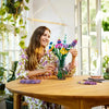 A woman holds a bouquet of colorful flowers in a glass vase on a wooden table.