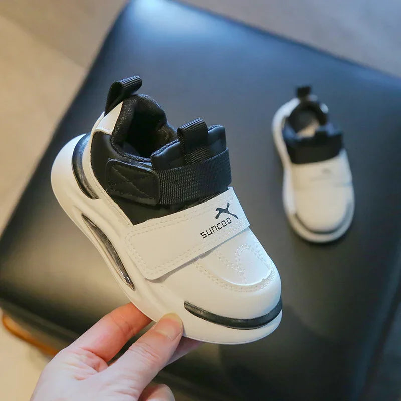 A hand holds a white and black kids' everyday sneaker with easy velcro straps on a dark surface.