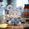 A fluffy bear plush charm with blue accents and a knitted scarf sits on a wooden surface.