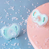 A soft, light blue silicone baby pacifier rests on a pink surface surrounded by scattered small white beads.