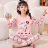 A young girl wearing a pink long-sleeved pajama set with a cat print and checkered pants sitting on a white couch.