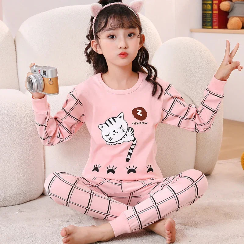 A young girl wearing a pink long-sleeved pajama set with a cat print and checkered pants sitting on a white couch.