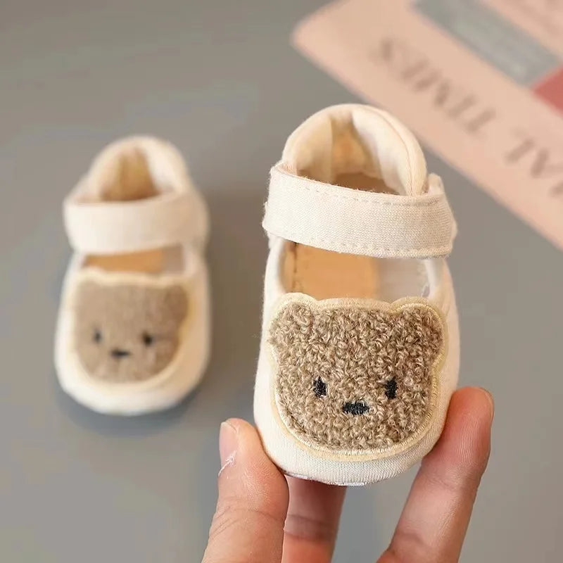 A pair of adorable bear soft sole first walker shoes for babies in cream color being held by a hand.