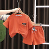 An orange boys' summer cotton t-shirt and shorts set displayed on a hanger against a dark curtain.