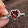 A close-up of a heart-shaped ring featuring a large red gemstone surrounded by pave diamonds being held by a finger.