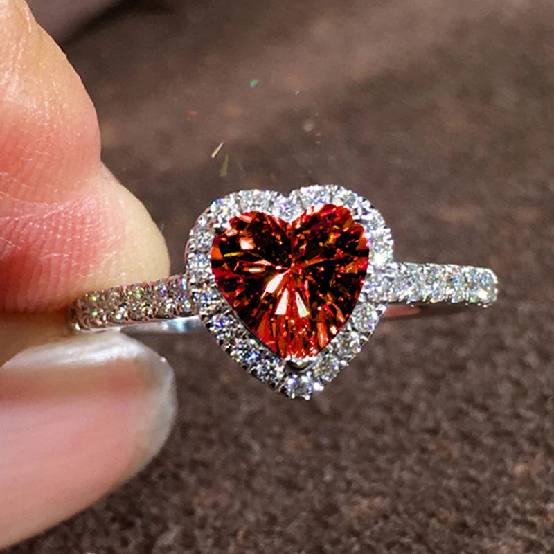 A close-up of a heart-shaped ring featuring a large red gemstone surrounded by pave diamonds being held by a finger.