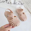 A hand holds a pair of light pink girls' gladiator jelly sandals with woven straps and non-slip soles.