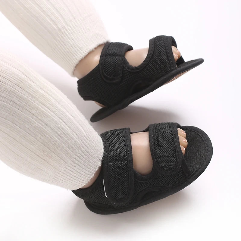 Black breathable first walker sandals for baby with hook-and-loop closure shown on a foot wearing a white sock.