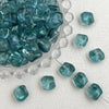 A clear glass bowl filled with irregular square-shaped teal blue beads and loose beads on a white surface.