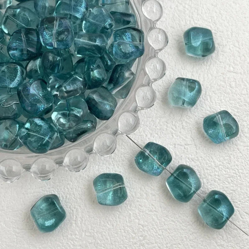 A clear glass bowl filled with irregular square-shaped teal blue beads and loose beads on a white surface.