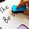 A hand is holding a bright blue magnetic dry erase eraser on a white board, writing the word Be so.
