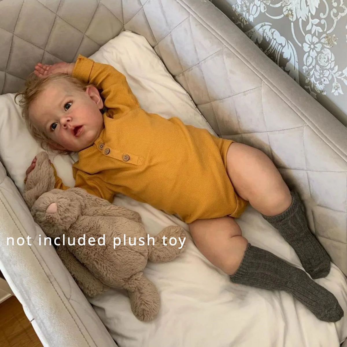 A baby girl lies in a white crib bedding, wearing a yellow outfit and socks, with a plush toy beside her.