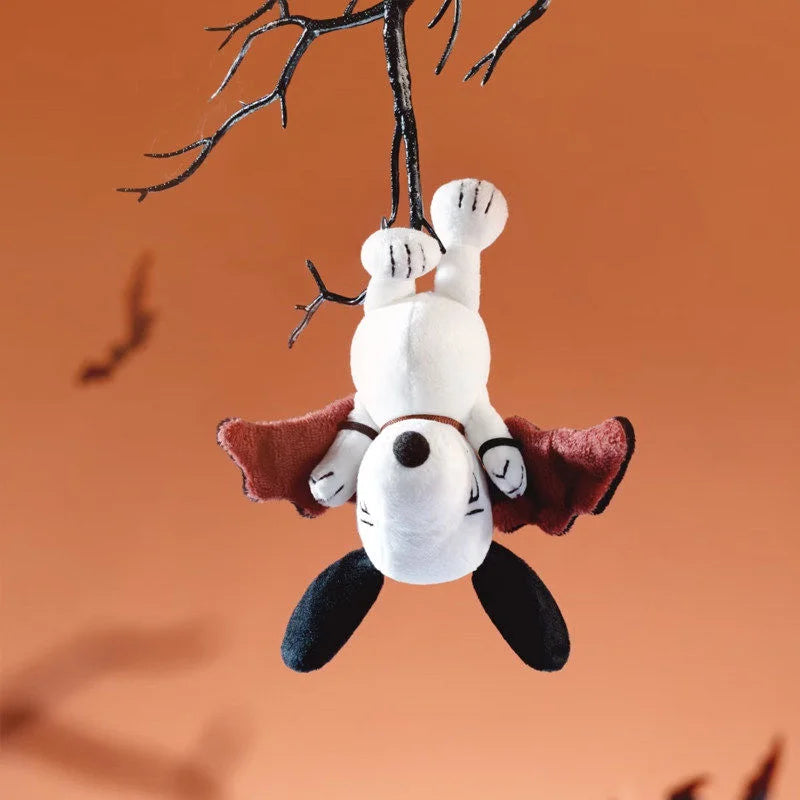 A white Snoopy collectible figure with brown ears and a red cape is suspended from bare branches against an orange background
