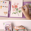 A hand is painting a floral watercolor illustration in an open coloring book, next to other art supplies.