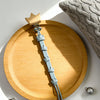 A silver baby pacifier clip with a burlap flower design rests on a wooden plate next to a knitted gray cushion.