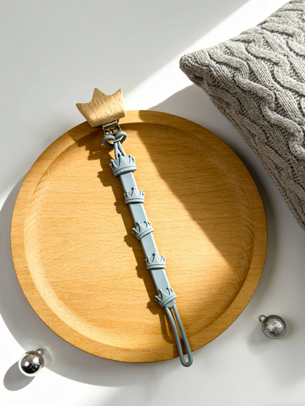A silver baby pacifier clip with a burlap flower design rests on a wooden plate next to a knitted gray cushion.