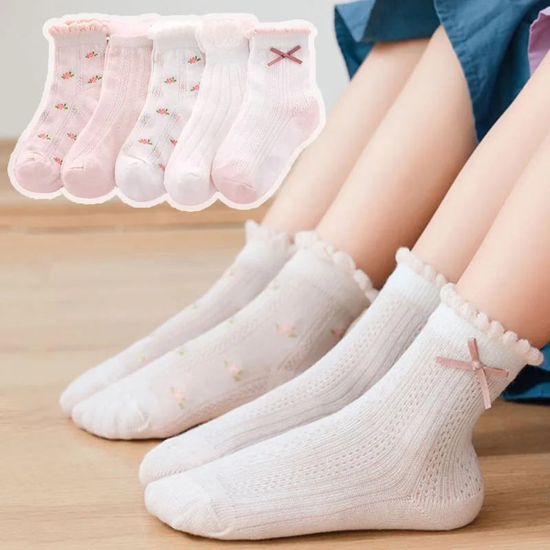 A stack of five pink floral cotton kids socks and three white knitted socks are displayed on a wooden surface.