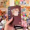 A hand holds a brown sticker book with a dark purple ribbon, featuring retro designs and colorful stickers.