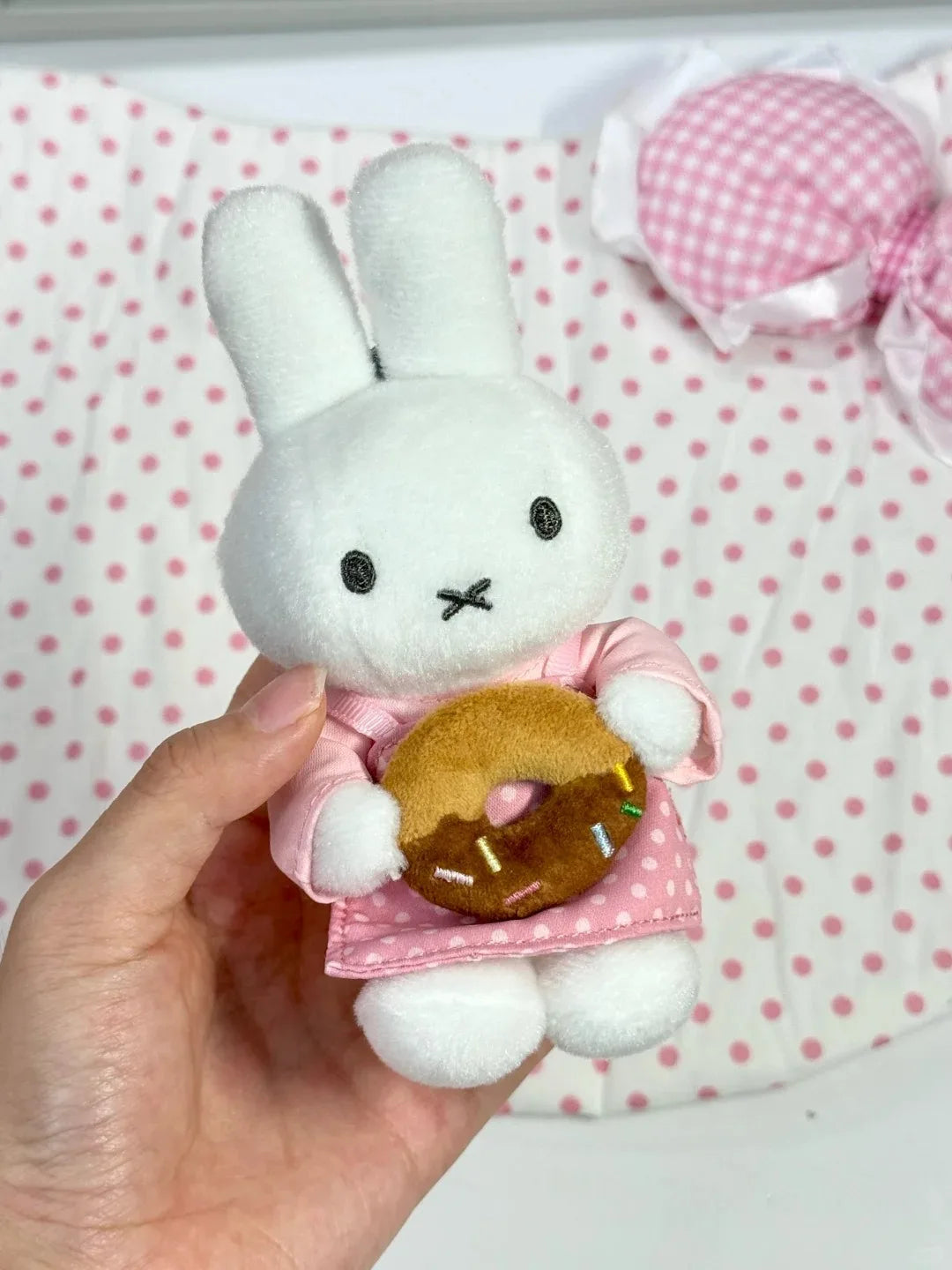 A white bunny plush toy with pink clothing and a golden donut charm is held in a hand.