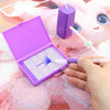 A hand is using a white pen to apply glitter onto a purple diamond painting canvas inside an open purple box.