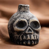 A ceramic Aztec skull whistle with dark gray and white paint resting on brown fabric.
